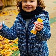Alma a rejoint le concours — aidez-le/la à gagner de superbes lots ! child, curly_hair, blue_jacket, bubble_wand, bubble, outdoor, autumn, leaves, path, smiling, happy, scarf, playful, person, fall, young_child, nature, jacket, toy, daylight