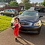 black_car, box, car, child, curly_hair, driveway, grass, house, infiniti, outdoor, parking, person, red_dress, shadow, sidewalk, sneakers, sunlight, tablet, trees, walking