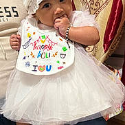 Avika joined the competition — help win amazing prizes! baby, child, infant, white_dress, hat, bib, socks, bracelet, hand, person, indoor, curtains, pattern, floor, lap, cute, festive, happy, colorful, portrait