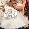 baby, child, infant, white_dress, hat, bib, socks, bracelet, hand, person, indoor, curtains, pattern, floor, lap, cute, festive, happy, colorful, portrait