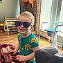 Walker is registered to the contest to win money with this photo: toddler, child, sunglasses, banana_print, onesie, smiling, blanket, indoor, living_room, wood_floor, furniture, toy_storage, playroom, happy, cute, sunlight, window, chair, face, person
