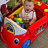 baby, bib, carpet, child, christmas, colorful, cute, decorations, festive, happy, holiday, indoor, infant, person, play, red, sitting, smiling, steering_wheel, toy_car