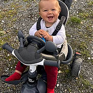 Aléssio participe au concours pour gagner de l'argent avec cette photo : baby, child, tricycle, stroller, outdoor, smiling, happy, red_socks, white_top, toy, play, ground, grass, rocks, person, seat, handlebars, wheels, hands, face
