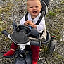 baby, child, tricycle, stroller, outdoor, smiling, happy, red_socks, white_top, toy, play, ground, grass, rocks, person, seat, handlebars, wheels, hands, face