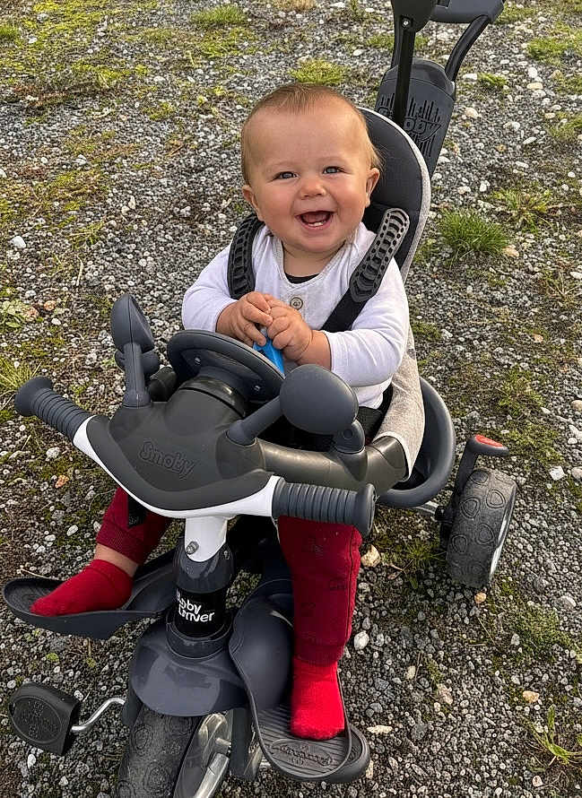 Aléssio participe au concours pour gagner de l'argent avec cette photo : baby, child, tricycle, stroller, outdoor, smiling, happy, red_socks, white_top, toy, play, ground, grass, rocks, person, seat, handlebars, wheels, hands, face