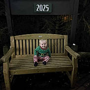 Archie is registered to the contest to win money with this photo: baby, bench, wood, forest, night, sign, clothing, child, smile, person, outdoor, holiday, pajamas, striped_pants, green_top, seat, wooden_bench, dark, trees, happy