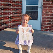 Lakelyn joined the competition — help win amazing prizes! toddler, child, girl, dress, white_tights, white_shoes, holding_artwork, bunny_artwork, brick_wall, concrete_porch, door, outdoor, sunlight, shadow, happy, smiling, standing, hair_blowing, front_porch, casual