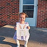 toddler, child, girl, dress, white_tights, white_shoes, holding_artwork, bunny_artwork, brick_wall, concrete_porch, door, outdoor, sunlight, shadow, happy, smiling, standing, hair_blowing, front_porch, casual