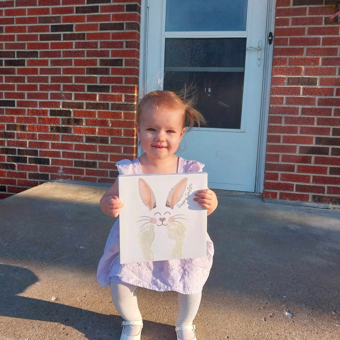 Lakelyn joined the competition — help win amazing prizes! toddler, child, girl, dress, white_tights, white_shoes, holding_artwork, bunny_artwork, brick_wall, concrete_porch, door, outdoor, sunlight, shadow, happy, smiling, standing, hair_blowing, front_porch, casual