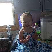 Lakelyn is registered to the contest to win money with this photo: toddler, child, smiling, clapping, dress, blue_and_white, kitchen, counter, window, dish_soap, soda_bottle, appliance, cabinet, natural_light, happy, person, indoor, hair_bow, barefoot, portrait