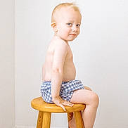 Lip a rejoint le concours — aidez-le/la à gagner de superbes lots ! toddler, child, boy, sitting, wooden_stool, shorts, bare_chest, light_hair, blue_eyes, carpet, indoor, minimalist, smile, pose, furniture, person, portrait, skin, room, wall