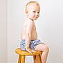 toddler, child, boy, sitting, wooden_stool, shorts, bare_chest, light_hair, blue_eyes, carpet, indoor, minimalist, smile, pose, furniture, person, portrait, skin, room, wall