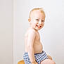toddler, child, smiling, sitting, stool, blond_hair, bare_chest, blue_shorts, checkered, indoor, white_wall, wooden_furniture, happy, portrait, cute, young_child, skin, person, innocence, playful