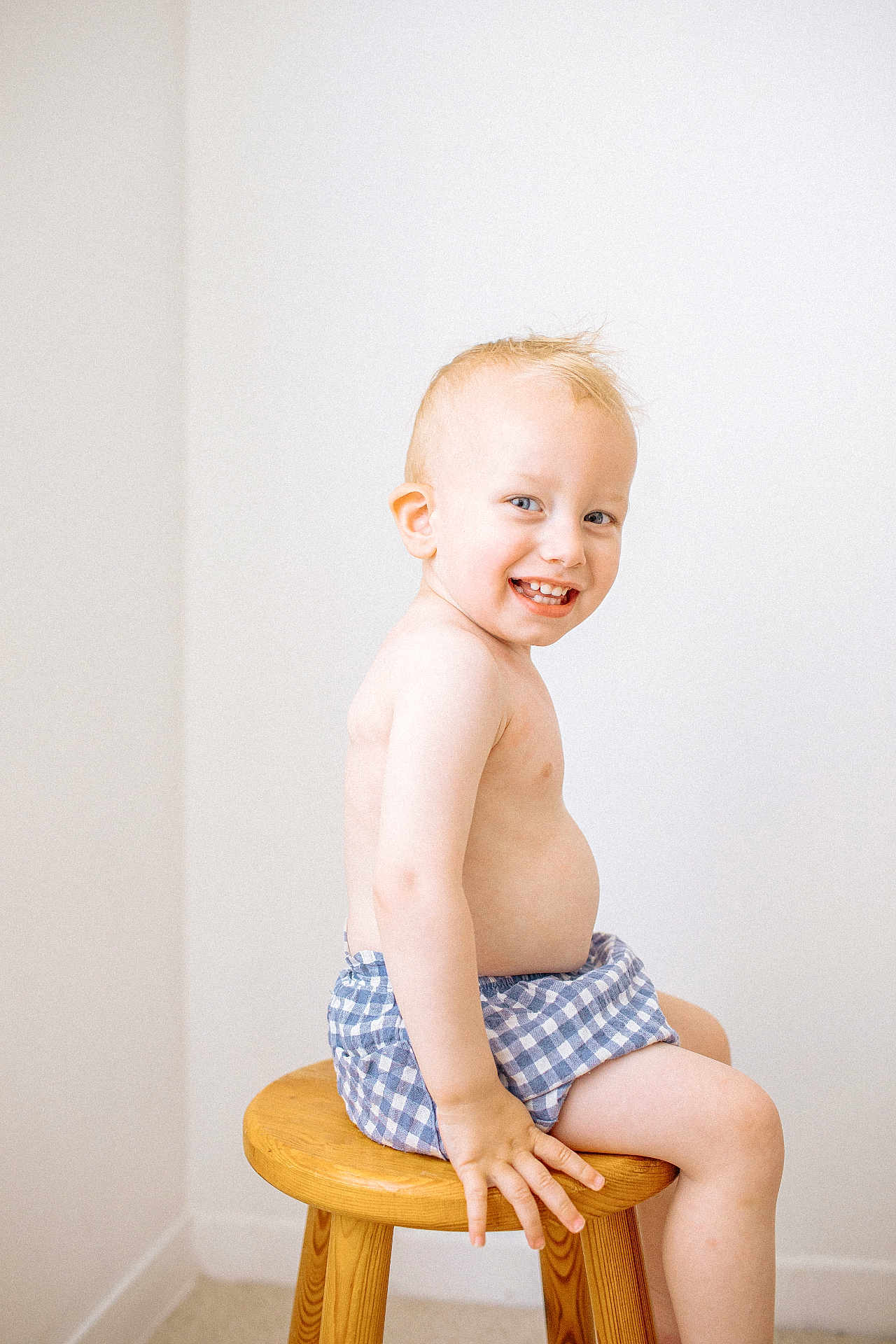 Lip participe au concours pour gagner de l'argent avec cette photo : toddler, child, smiling, sitting, stool, blond_hair, bare_chest, blue_shorts, checkered, indoor, white_wall, wooden_furniture, happy, portrait, cute, young_child, skin, person, innocence, playful