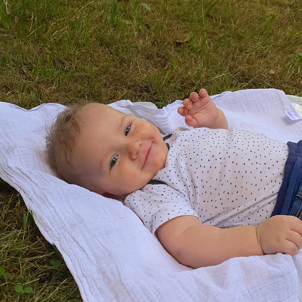 Baptiste participe au concours pour gagner de l'argent avec cette photo : baby, blanket, casual_clothing, child, cute, daylight, grass, greenery, happy, infant, lying_down, nature, outdoor, person, playful, portrait, relaxing, smiling, trees, white_blanket