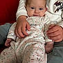 baby, child, person, infant, couch, clothing, sweatshirt, onesie, hand, face, indoor, red_couch, seated, comfort, cute, portrait, young_child, holding, casual, expression