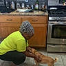 dog, person, kitchen, indoor, crouching, petting, happy, animal, short_hair, brown_dog, tail, floor, stove, oven, countertop, cabinets, sink, bottle, home, cooking