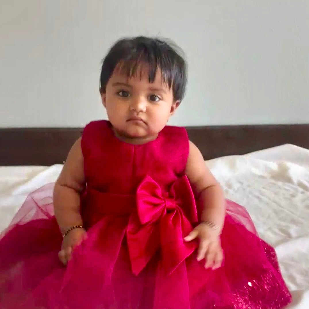 Dhanvi is registered to the contest to win money with this photo: baby, child, red_dress, bow, sitting, indoors, bed, white_bedspread, serious_expression, short_hair, bracelet, portrait, cute, infant, young_child, people, fashion, celebration, tulle, sparkle