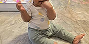 Julie Kay is registered to the contest to win money with this photo: baby, child, indoor, floor, marble_floor, sitting, toys, playpen, person, curly_hair, double_buns, casual_clothing, pants, tshirt, hand_in_mouth, bright, home, cute, curious, young_child