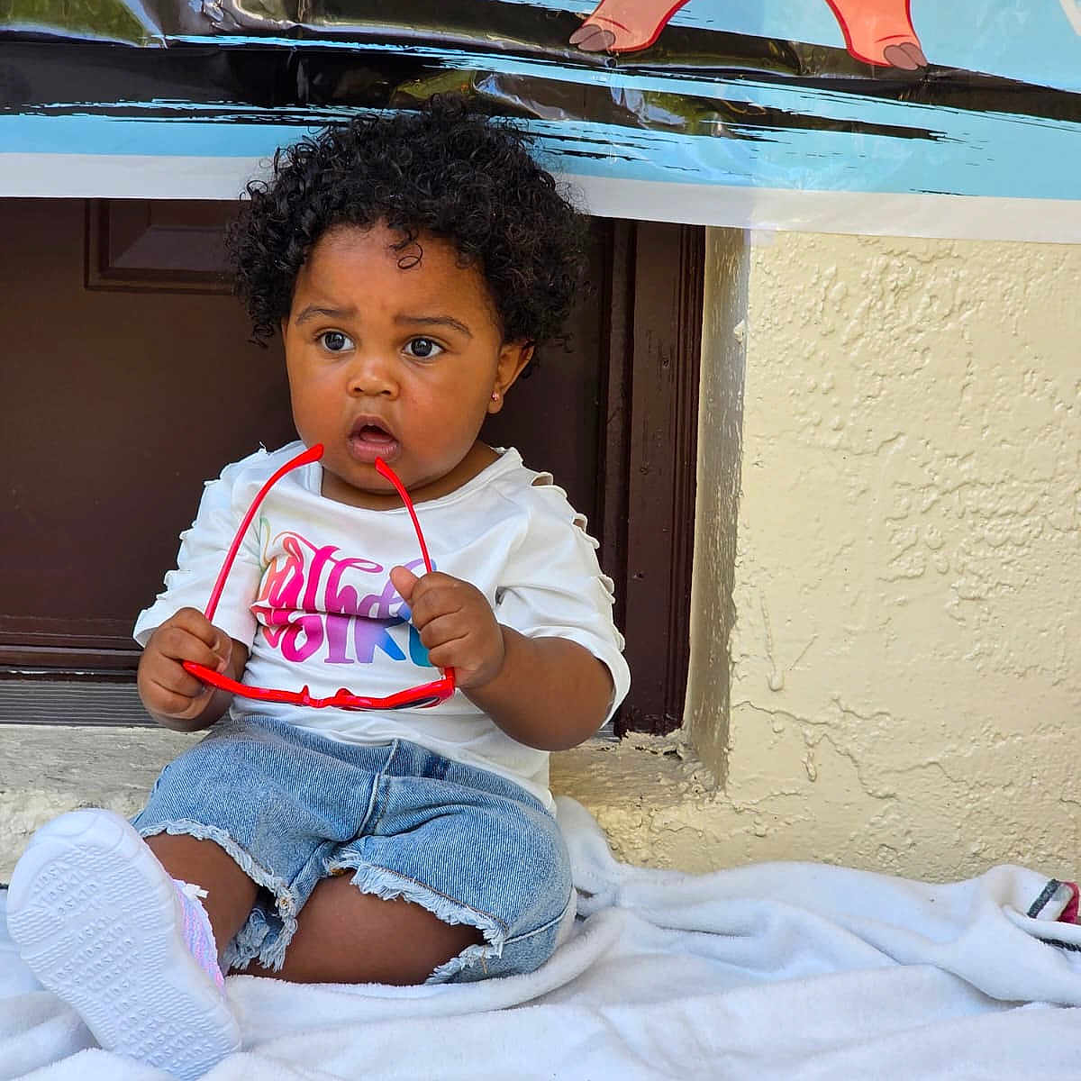 Julie Kay joined the competition — help win amazing prizes! banner, cartoon, casual, child, clothing, curly_hair, cute, denim_shorts, expression, footwear, outdoor, person, pig, playful, sitting, sunglasses, texture, toddler, wall, white_blanket