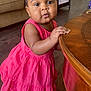 Julie Kay is registered to the contest to win money with this photo: toddler, child, pink_dress, standing, table, wooden_table, pigtails, hair_ties, living_room, couch, cushions, playpen, floor, barefoot, curious, indoors, furniture, person, portrait, home