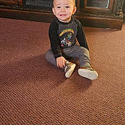 Wyatt is registered to the contest to win money with this photo: toddler, child, carpet, indoor, cabinet, wooden_furniture, glass_door, smile, sitting, clothing, shoes, person, face, toy_graphic, floor, home, room, young_child, cute, casual