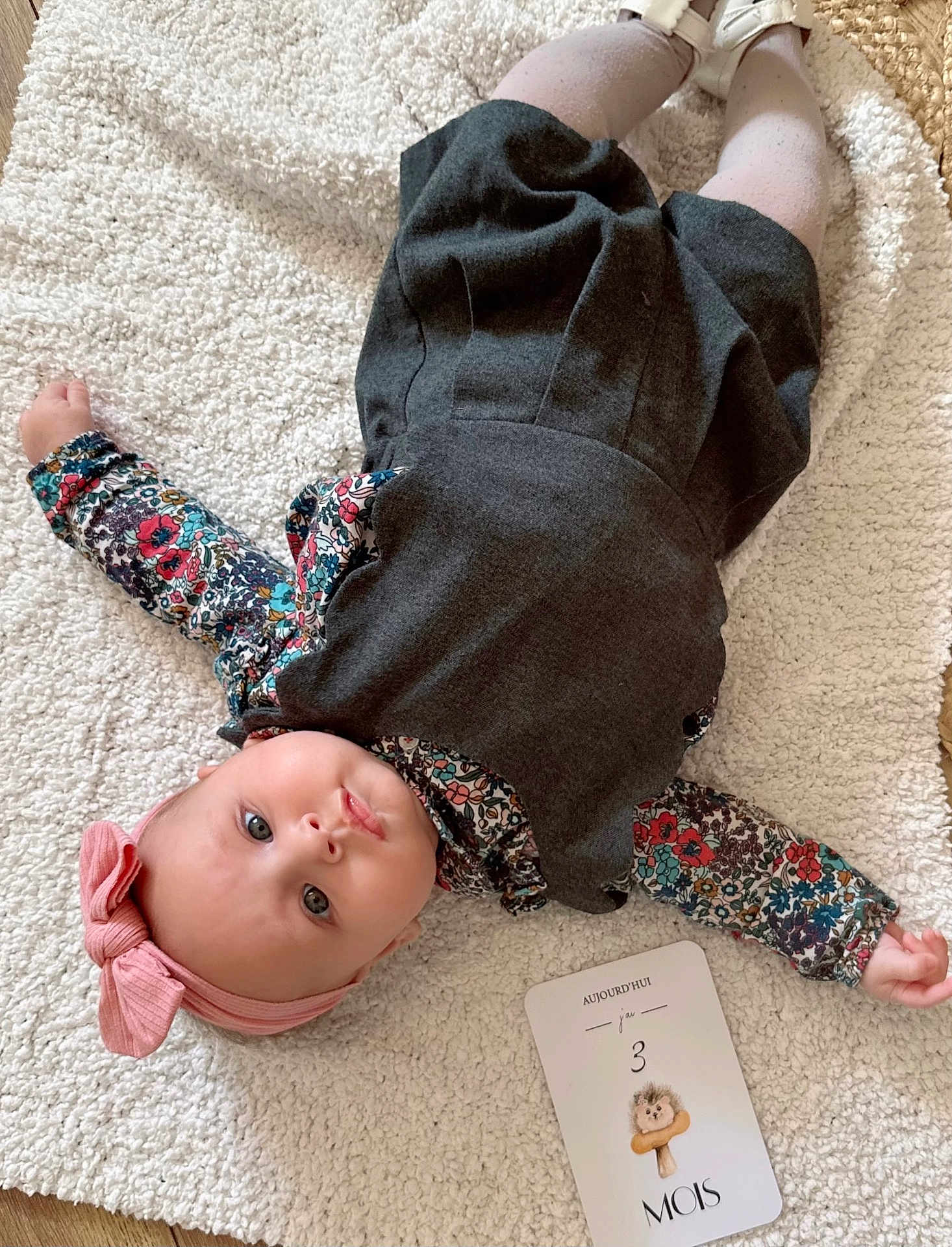 Victoire a rejoint le concours — aidez-le/la à gagner de superbes lots ! baby, infant, child, blanket, headband, bow, dress, floral_clothing, milestone_card, floor, soft_texture, cute, lying_down, indoors, portrait, pink, white, grey, wood_floor, curious