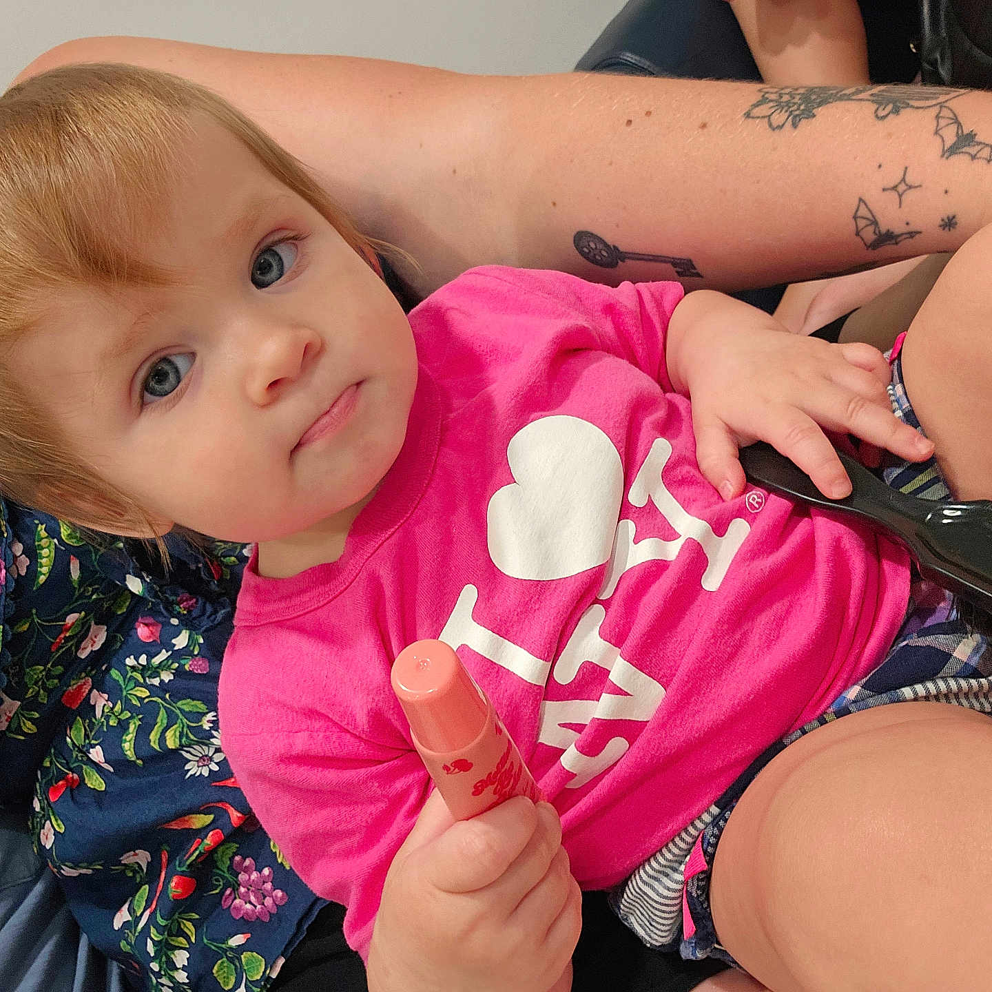 Olive is registered to the contest to win money with this photo: toddler, child, person, pink_shirt, lip_balm, hairbrush, tattoos, arm, floral_clothing, indoor, seated, blue_eyes, short_hair, skin, casual, holding_object, curious_expression, adult, closeup, candid