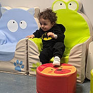Adam joined the competition — help win amazing prizes! batman_hoodie, blue_cat_chair, child, colorful, curly_hair, cute, floor, footwear, frog_chair, furniture, indoor, lion_face, play_area, red_stool, seated, smiling, toddler, wall, white_shoes, young_child