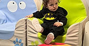 Adam is registered to the contest to win money with this photo: batman_hoodie, blue_chair, casual, cat_chair, child, clothing, colorful, comfortable, curly_hair, floor, footwear, frog_chair, green_chair, indoor, lion_face, playful, red_stool, sitting, toddler, young_child