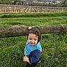 child, toddler, curly_hair, blue_jacket, wooden_fence, grass, vineyard, outdoor, nature, greenery, field, rural, playful, person, young_child, daylight, scenic, landscape, casual_clothing, smiling