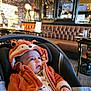 baby, child, hoodie, orange_clothing, car_seat, indoor, cafe, leather_seat, curious, cozy, warm_lighting, face, expression, cute, young_child, casual_clothing, seated, winter_wear, portrait, infant