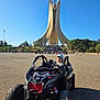 child, toddler, toy_car, outdoor, monument, blue_sky, daylight, vehicle, play, fun, people, road, park, cap, smile, trees, architecture, wheel, sunny, recreation