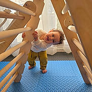 Adam a rejoint le concours — aidez-le/la à gagner de superbes lots ! toddler, child, playing, wooden_frame, climbing_toy, barefoot, indoor, blue_mat, green_ball, curtains, natural_light, happy, smiling, casual_clothing, yellow_pants, white_sweatshirt, curly_hair, floor_mat, home, fun