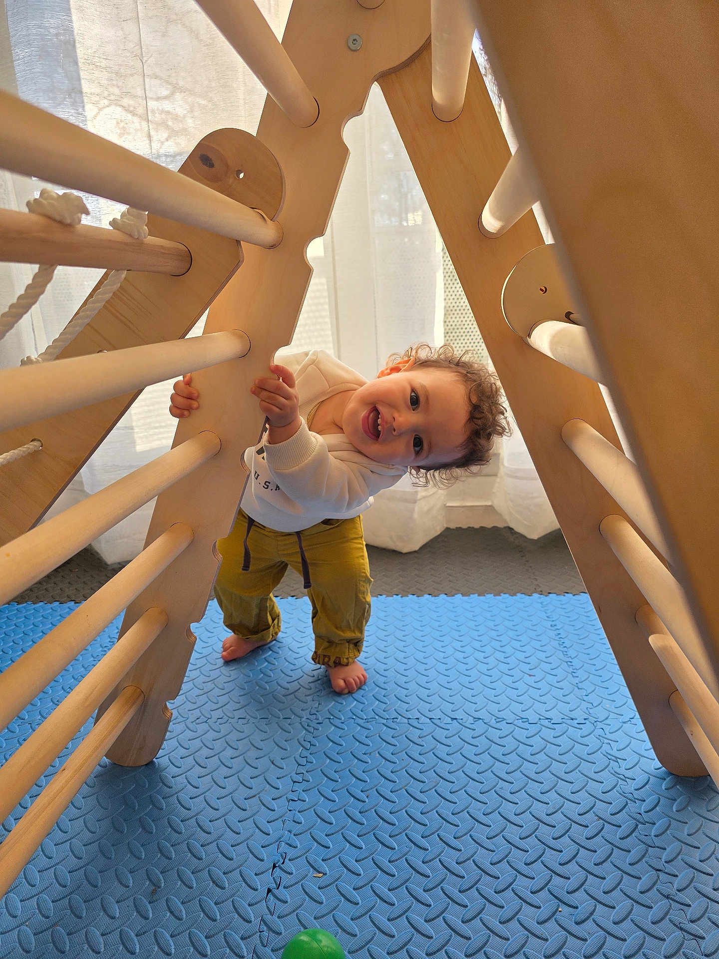 Adam a rejoint le concours — aidez-le/la à gagner de superbes lots ! toddler, child, playing, wooden_frame, climbing_toy, barefoot, indoor, blue_mat, green_ball, curtains, natural_light, happy, smiling, casual_clothing, yellow_pants, white_sweatshirt, curly_hair, floor_mat, home, fun