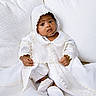 baby, infant, child, white_clothing, bonnet, christening_gown, cushion, fabric, portrait, sitting, cute, indoors, soft_lighting, closeup, face, hands, footwear, bracelet, serene, curious