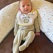 Sandro participe au concours pour gagner de l'argent avec cette photo : baby, infant, onesie, bib, pillow, patterned_pillow, brown_background, face, smile, sitting, curious, soft_clothing, head, indoor, cute, child, person, relaxed, baby_bib, sleepwear