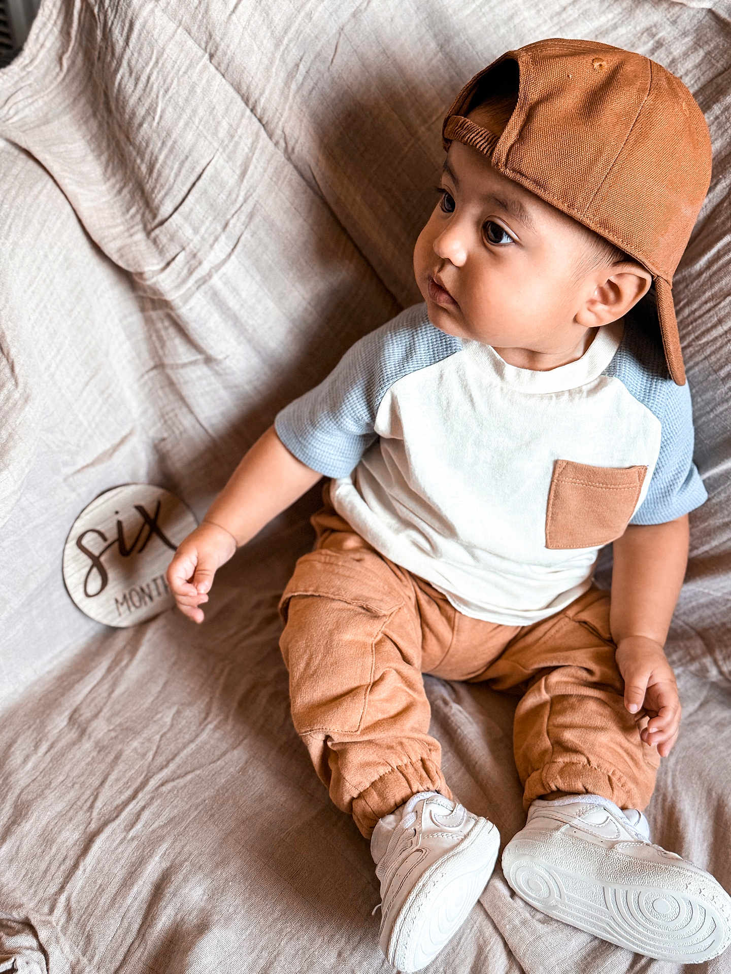 Juan is registered to the contest to win money with this photo: baby, child, infant, cap, brown_clothing, white_shoes, sitting, blanket, milestone_sign, six_months, curious, side_view, indoor, portrait, casual_clothes, cute, soft_lighting, cozy, young_child, fabric