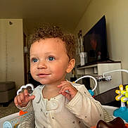 Isabel joined the competition — help win amazing prizes! child, toddler, curly_hair, blue_eyes, play_chair, toy, indoor, smile, cute, baby_clothes, baby, face, person, living_room, furniture, television, curiosity, playtime, happy, expression
