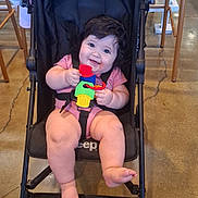 Maria is registered to the contest to win money with this photo: baby, child, stroller, smiling, happy, portrait, toy, colorful_toy, pink_clothing, seated, infant, face, eyes, hair, indoor, restaurant, chair, floor, wheels, family_outing