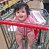 Maria is registered to the contest to win money with this photo: baby, child, infant, shopping_cart, supermarket, grocery_store, aisle, portrait, smiling, happy, sitting, pink_clothing, blanket, bare_feet, face, dark_hair, indoor, shopping, retail, cart_handle