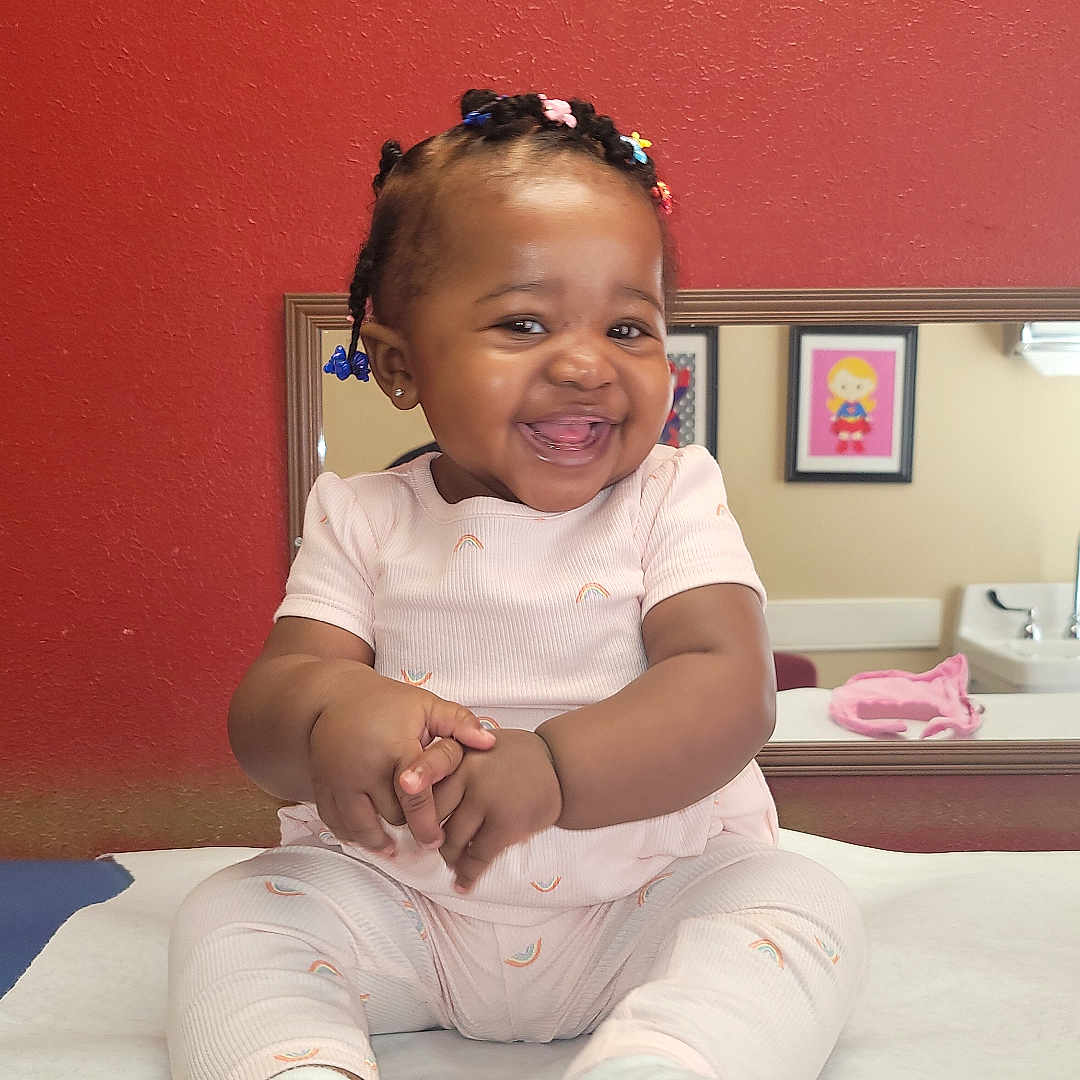 Kueen is registered to the contest to win money with this photo: baby, child, clapping, colorful_hair_beads, cute, framed_art, furniture, happy, indoor, infant, medical_table, mirror, pink_outfit, portrait, red_wall, room, sitting, skin, smiling, white_socks