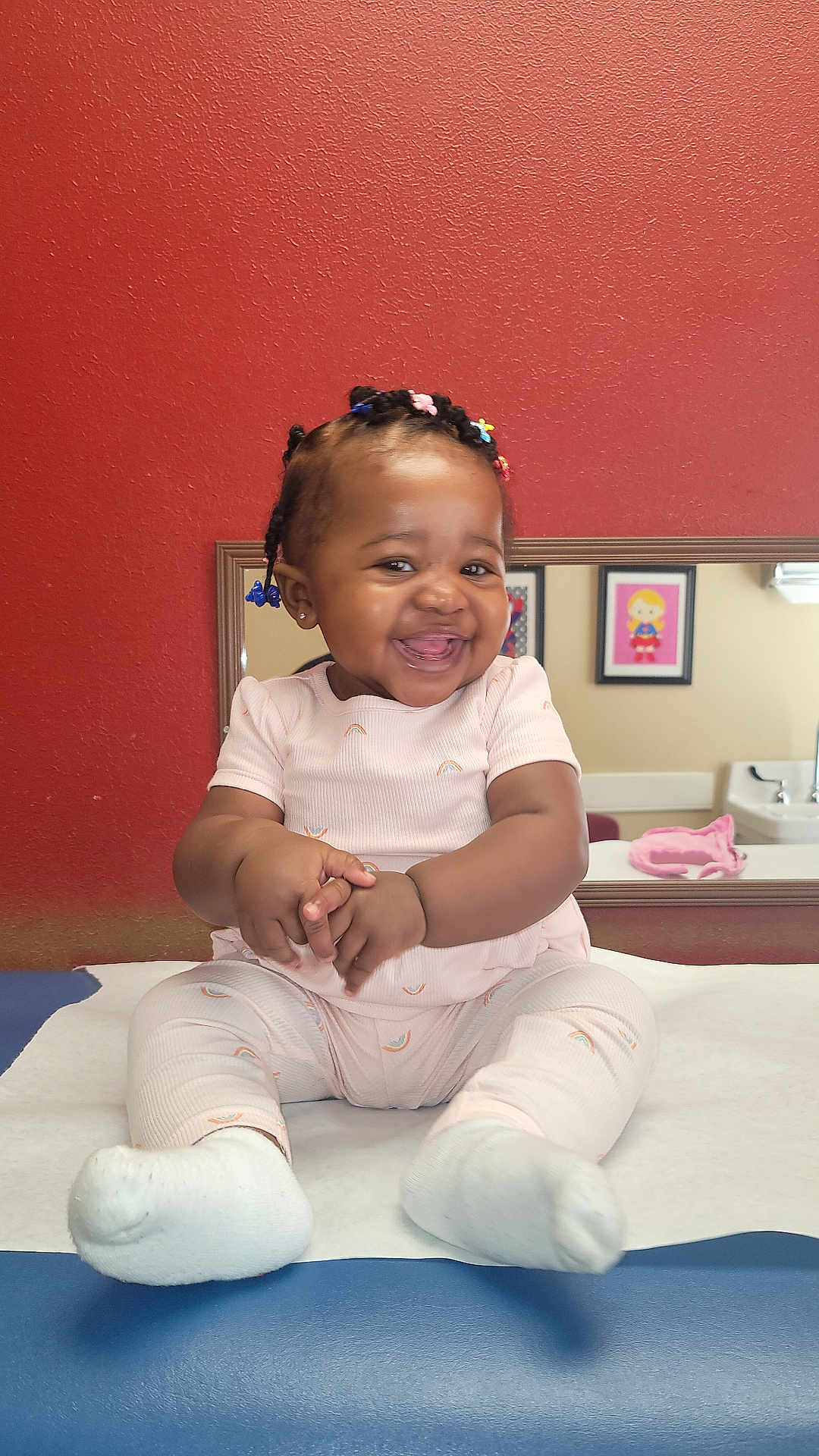 Kueen is registered to the contest to win money with this photo: baby, child, smiling, happy, sitting, clapping, medical_table, indoor, red_wall, mirror, framed_art, colorful_hair_beads, white_socks, pink_outfit, cute, portrait, infant, skin, room, furniture