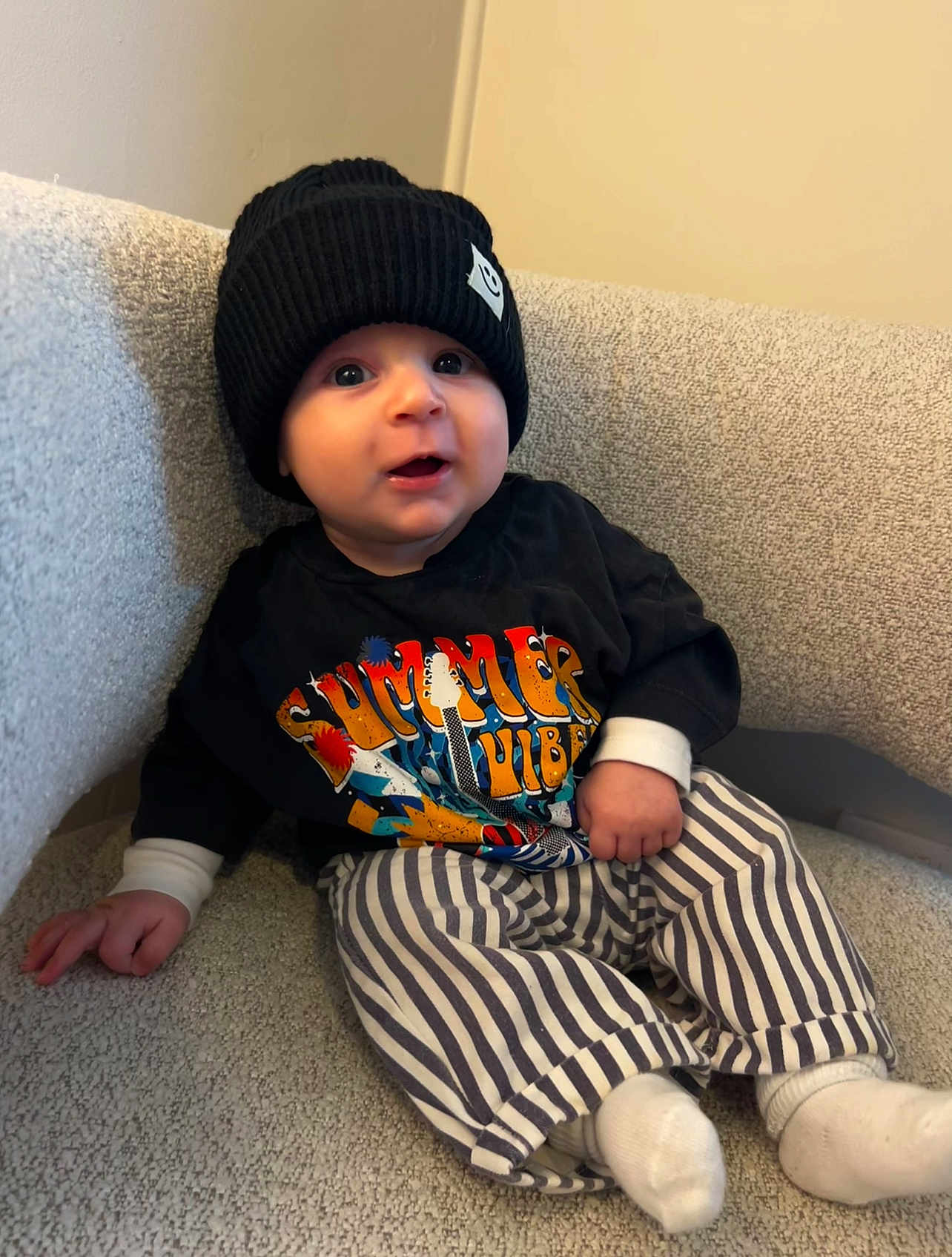 Cohen is registered to the contest to win money with this photo: baby, child, beanie, hat, striped_pants, socks, couch, indoor, portrait, cute, smiling, black_clothing, white_socks, infant, seated, happy, face, person, cozy, relaxed