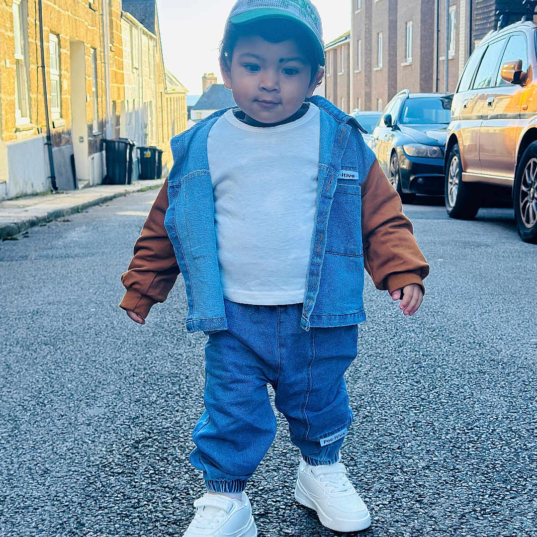 Apaar is registered to the contest to win money with this photo: asphalt, buildings, cap, casual_clothing, child, confidence, cool, daytime, denim_jacket, fashion, outdoor, person, sidewalk, small_person, street, toddler, urban, walking, white_shoes, young_child