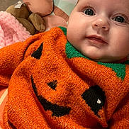 Esme is registered to the contest to win money with this photo: baby, infant, orange_sweater, jack_o_lantern, cute, close_up, plush_toy, blanket, soft_texture, portrait, child, face, eyes, smile, cozy, indoors, knitwear, holiday_theme, happy, clothing