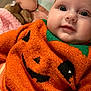 baby, infant, orange_sweater, jack_o_lantern, cute, close_up, plush_toy, blanket, soft_texture, portrait, child, face, eyes, smile, cozy, indoors, knitwear, holiday_theme, happy, clothing