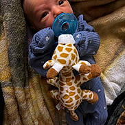 Carmella is registered to the contest to win money with this photo: baby, pacifier, plush_toy, giraffe, blanket, blue_clothing, infant, cute, soft_texture, cozy, sleepwear, child, newborn, resting, comfort, warm_colors, indoor, portrait, small_hands, cute_expression