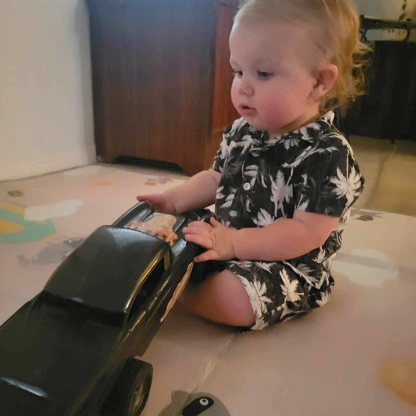 Lucius is registered to the contest to win money with this photo: toddler, child, toy_car, play_mat, indoor, curly_hair, black_clothing, white_clothing, animal_print, raccoon_illustration, wooden_furniture, window, soft_lighting, floor, candle, lamp, person, cute, playing, baby