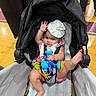 Malia joined the competition — help win amazing prizes! baby, stroller, toy, hand, foot, napkin_hat, floor, indoor, child, person, waving, colorful, playful, curious, seated, clothing, black_stroller, wooden_floor, people_legs, infant